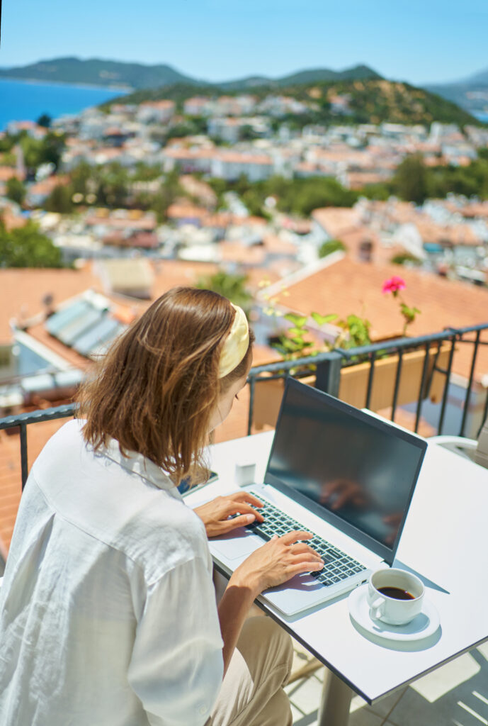 Rear view woman using laptop computer working typing emails browsing online over Mediterranean view from outside balcony travel hotel