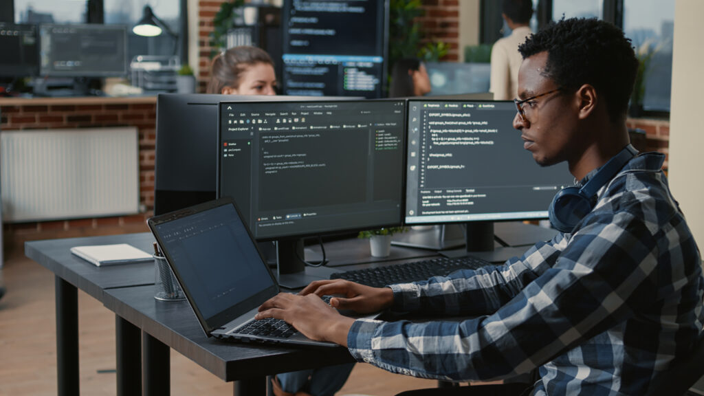 Portrait of tired programer rubbing eyes while typing on laptop sitting at desk with multiple screens parsing code in software agency. Coder losing focus while working on user interface.