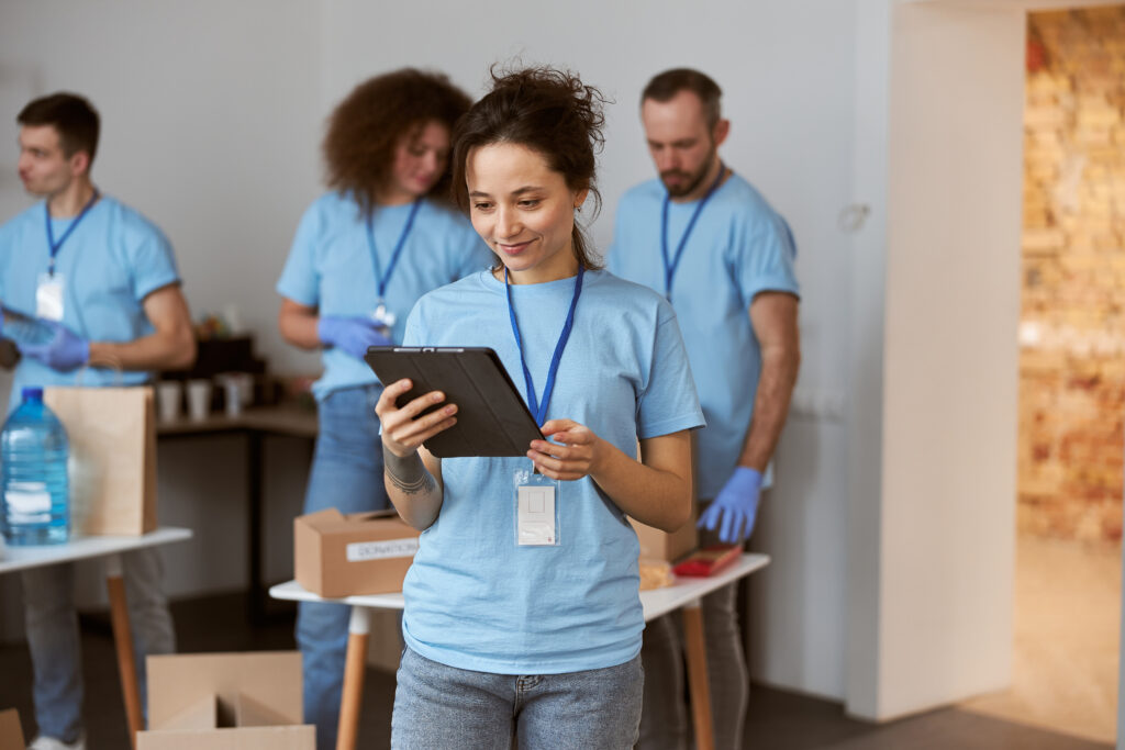Attractive young female volunteer in blue uniform using tablet pc and smiling while standing indoors. Team sorting, packing items in the background. Charitable foundation concept