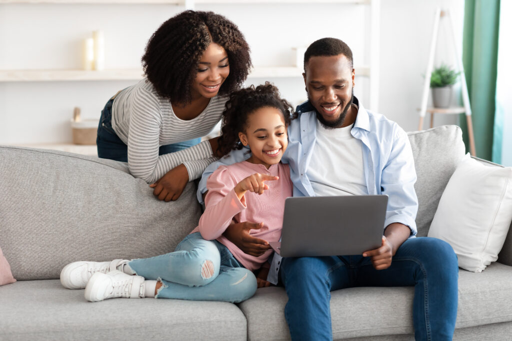 People And Technology Concept. Portrait of cheerful African American family of three using laptop, dad hugging daughter, teenage girl pointing at screen, showing photos or product in online webstore