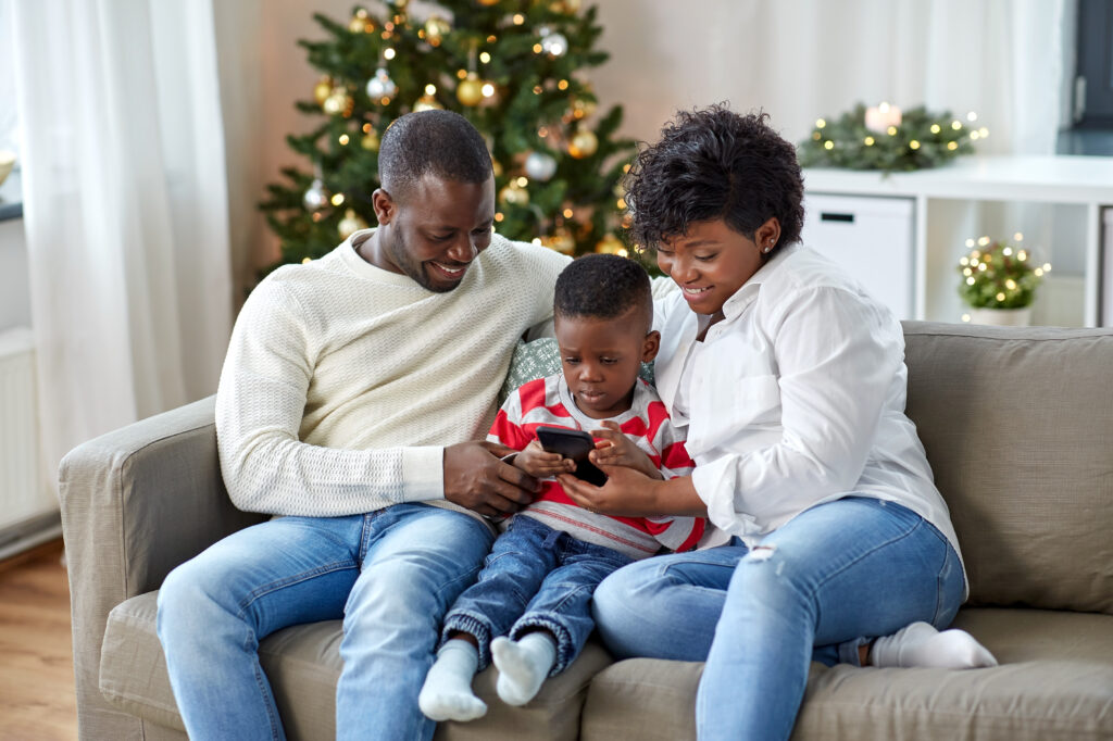 family, winter holidays and people concept - happy african american mother, father and little son with smartphone at home on christmas