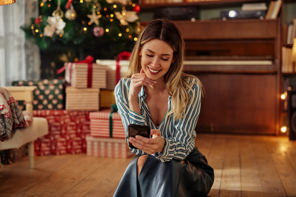 Positive excited young adult woman surfing net on smartphone while sitting on floor in decorated living room during Christmastime. Cheerful young woman chatting on social media.
